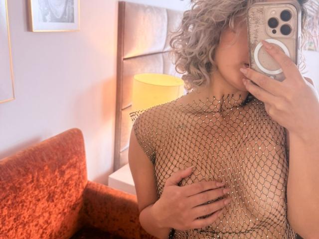 FloraSaenz with curly gray hair, wearing a gold mesh top, covering her tits with one hand, holding a phone with a ring grip, in a warm, orange-lit room.