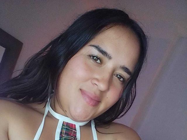 FunnyLesbians with long black hair, brown eyes, and a slight smile, wearing a white halter top with a red plaid pattern, showing her bare shoulders and upper chest.