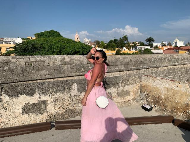 GalaMalkova with tan skin, black hair, and sunglasses, wearing a pink dress with a white pom-pom, standing on a stone rooftop, looking back, hand on head.