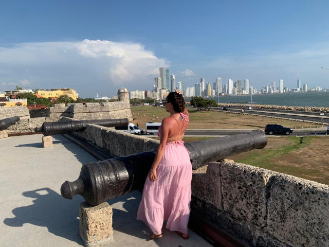 GalaMalkova se tient dos à la caméra, vêtue d'une robe rose à encolure américaine et d'un pantalon ample, ses cheveux bouclés attachés en chignon, près d'anciens canons, par une journée ensoleillée..