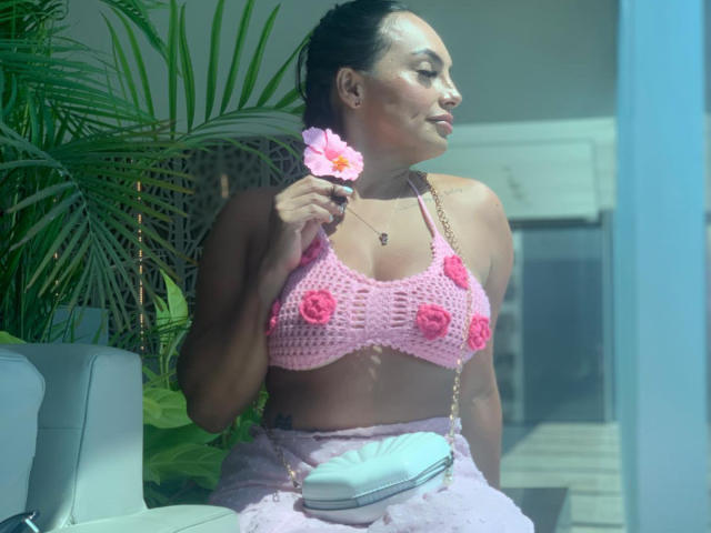 Gala Malkova mit gebräunter Haut, dunklem Haar zu einem Dutt gebunden, einem rosa Häkeloberteil, der ihre Brüste zeigt, hält eine rosa Blume, trägt einen rosa Rock und eine weiße Handtasche, in einem sonnendurchfluteten Raum..