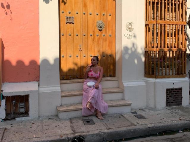 GalaMalkova mit rosa Brüsten in einem rosa BH und einem rosa Rock, sitzend auf Stufen, mit einer weißen Platte in der Hand, Holztür, rosa und weiße Wände, im Schatten, lässig, Tageslicht..