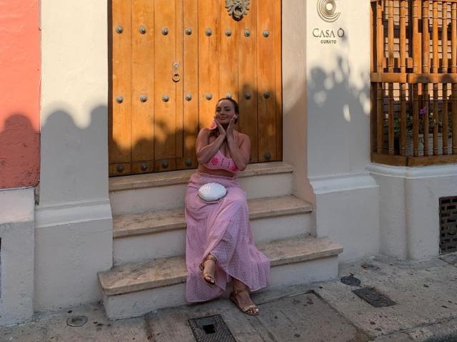 GalaMalkova sitting on stone steps, wearing a pink dress, hands near face, smiling, white purse on lap, wooden door behind, sunlight casting shadows, sandals on feet.