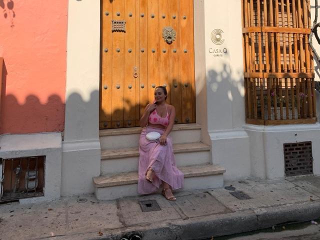 GalaMalkova met roze borsten en billen, gekleed in een roze jurk, zittend op trappen, met een witte kom in haar handen, een houten deur, zonneschaduwen, een roze muur, een witte muur en een houten hek..