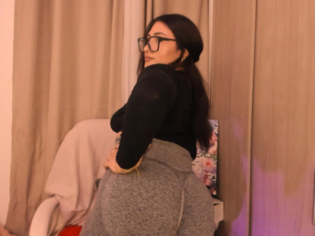 ElyRoss with dark hair, glasses, black top, and gray skirt, showing off her big ass, hands on hips, looking over shoulder in a room with beige curtains.