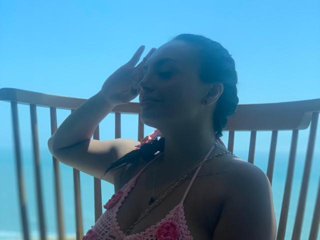 GalaMalkova with braided hair, pink lace top showing her tits, sunlit, hand on forehead, necklace, clear blue sky, wooden balcony, sea in background, subtle tattoo on left shoulder.
