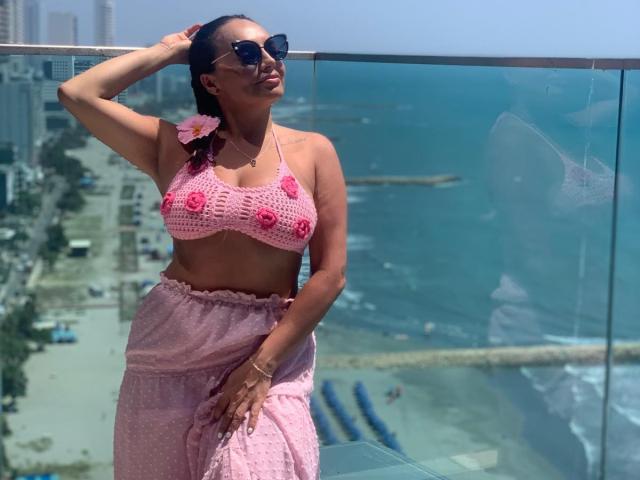 GalaMalkova met donkere huid, draagt een roze gebreide top met rozenversieringen, een roze rok en een zonnebril, en staat bij een glazen balkon, met haar hand in haar haar, een bloem in haar haar, met uitzicht op de oceaan..