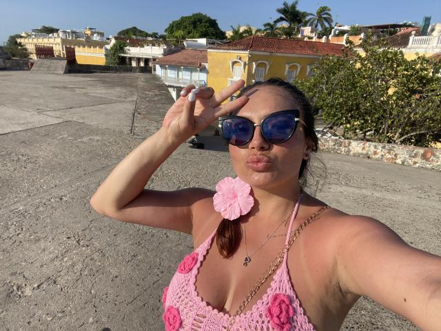 GalaMalkova com óculos de sol azuis, blusa de crochê rosa que mostra os seios, flor rosa no peito, lábios curvados, cabelo castanho em rabo de cavalo, telhado ensolarado, segurando o cabelo para cima, céu azul claro..