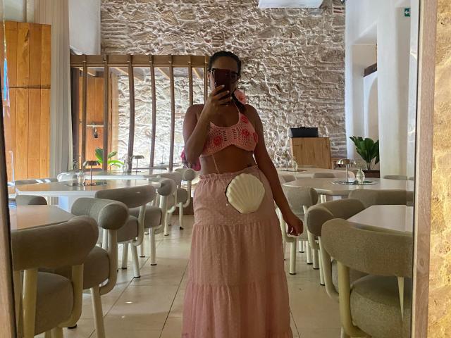 GalaMalkova está de pie en una cafetería, vestida con un corpiño de encaje rosa y pequeños senos, y una falda rosa, casi transparente, sosteniendo un teléfono frente a ella, con un abanico blanco..