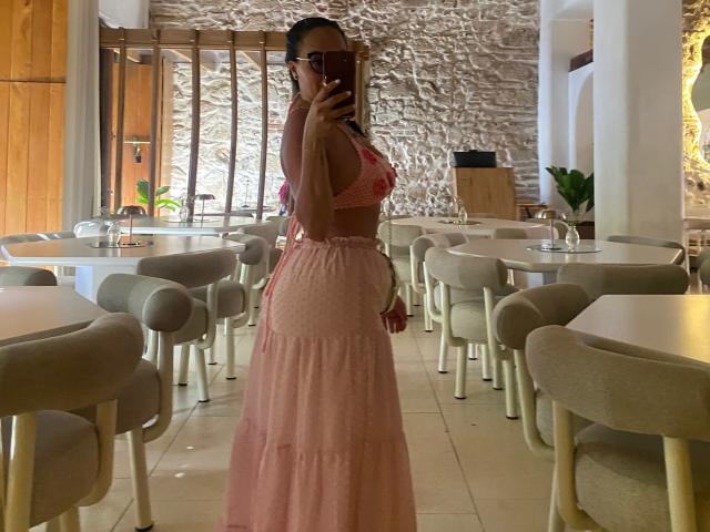 GalaMalkova, avec une peau bronzée, porte un soutien-gorge rose à motifs et une longue jupe rose, elle tient un téléphone, montre ses fesses, et se tient dans un café moderne avec des chaises grises et un mur en pierre..
