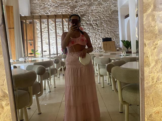 GalaMalkova met donkere huid, een roze gebreide top die haar borsten laat zien, een roze rok met hoge taille, die een witte clutch vasthoudt, staat op een spiegelzelfportret, in een restaurant met grijze stoelen..