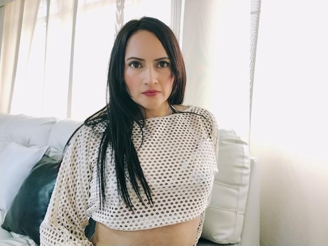 EmhilyDiaz, avec de longs cheveux noirs, portant un haut en maille blanche qui expose ses petites seins, se tient dans une pièce lumineuse, regardant directement l'appareil photo avec une expression sérieuse..