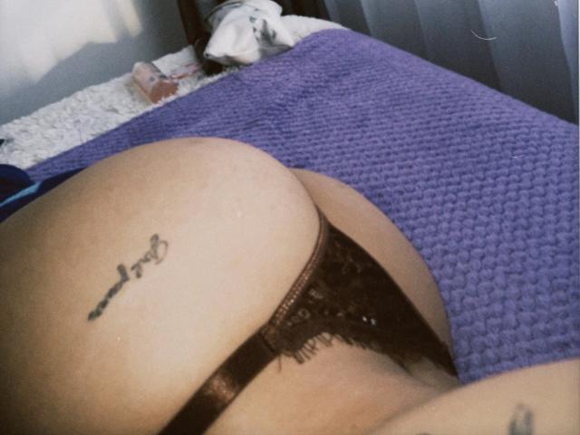 ThaliaAres' ass in black lace panties, showing a tattoo on her right buttock, lying on a purple textured blanket, with a white pillow in the background.