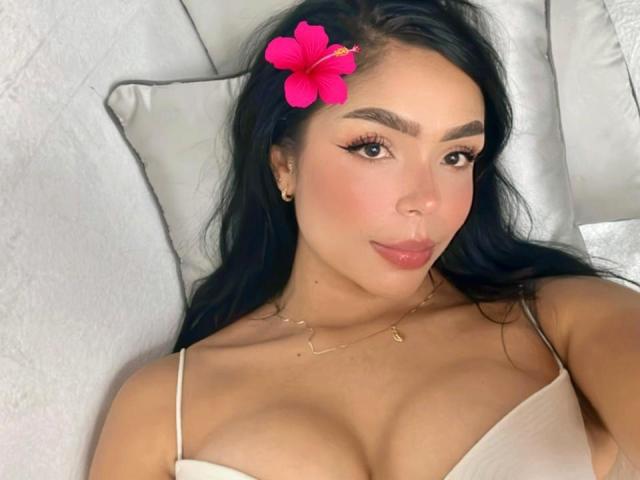 AnaGymez, com cabelo preto, uma flor de hibisco rosa no cabelo, uma blusa de alças finas branca, mostrando seus seios, deitada em travesseiros brancos, com maquiagem discreta, um colar de ouro e um leve sorriso..