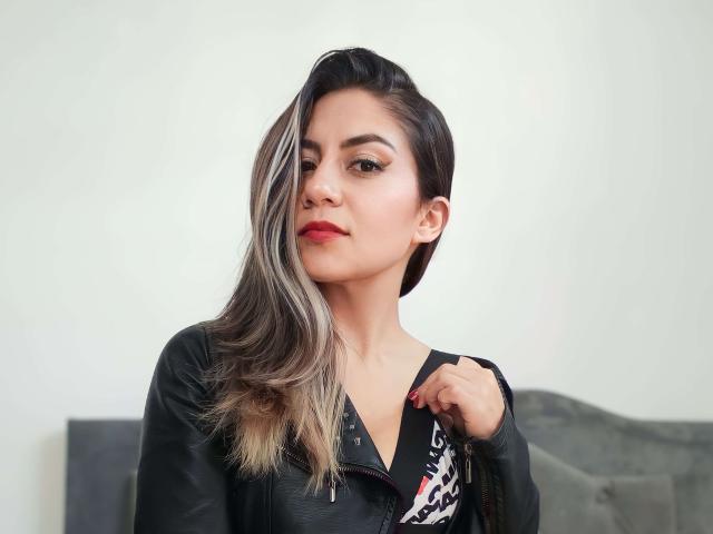ValeriaLoren con capelli corti, neri e biondi, indossa una maglietta nera, tiene una catena, ha il rossetto rosso e un leggero sorriso, e guarda direttamente la fotocamera..