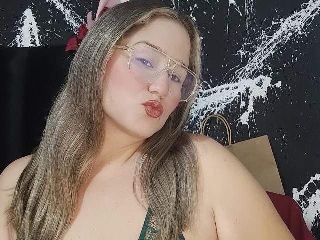 AlexaHotSeins with glasses, pouty lips, and blonde hair, wearing a green top, showing her tits, against a black and white splatter background.