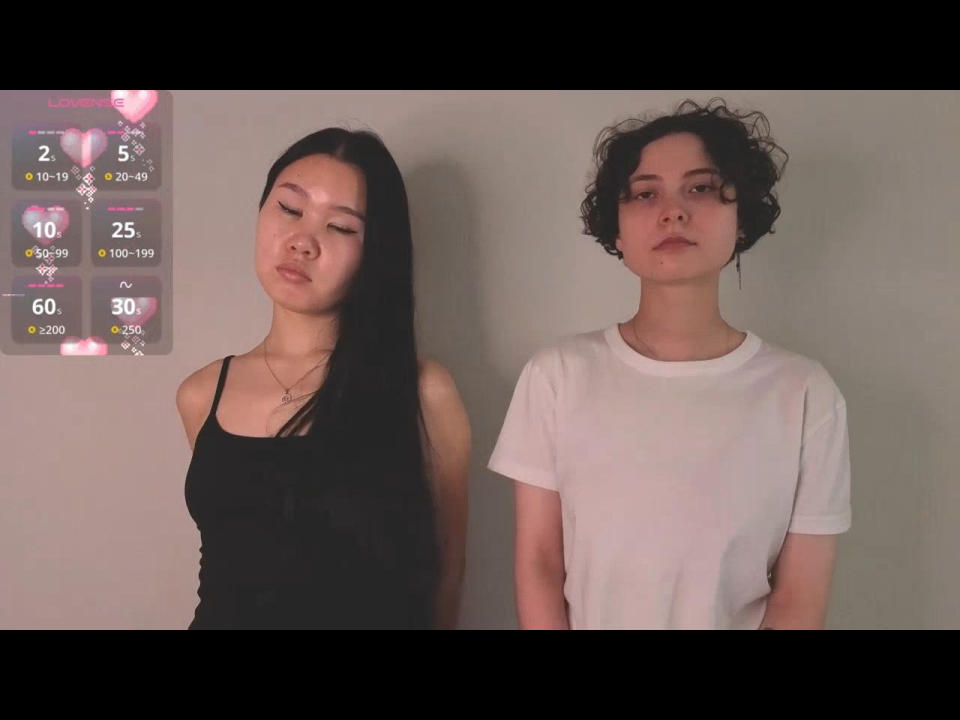 LatinaFeldker with long black hair, small tits, wearing a black tank top, standing next to curly-haired LatinaFeldker with white tee, both with serious faces.