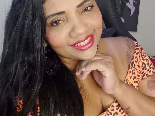 Samira, verführerisch mit langen, schwarzen Haaren, lächelt mit pinkem Lippenstift, trägt ein Top mit Leopardenmuster, zeigt ihre Brüste, hat ein Tattoo am Arm, trägt schwarze Creolen und eine niedliche Sommersprosse an der Nase..