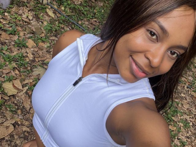 EmmaLatin with tan skin, brown eyes, and a smile, wearing a white zip-up top that shows her small tits, taking a selfie outdoors.