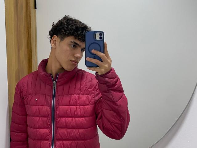 TheImperialMen with curly dark hair, wearing a pink puffy jacket, taking a mirror selfie with a blue phone, looking serious, standing in a white-walled room.