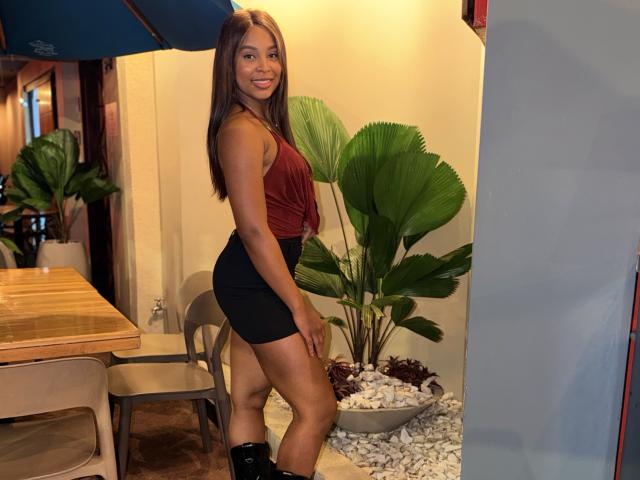 Emma, latina, com cabelos castanhos longos, vestindo uma blusa vermelha sem mangas e shorts pretos, sorrindo, em pé em um quarto, com uma planta verde ao lado, usando botas pretas, com curvas e com um bumbum pequeno..
