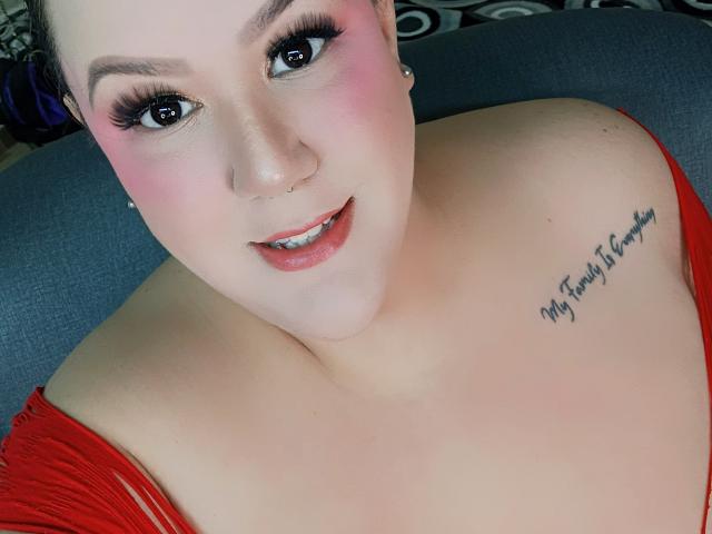 GiiannaMorrys with pink blush, black eyeliner, and red dress, showing tits with a "My Family is Everything" tattoo on her right boob, smiling at the camera.