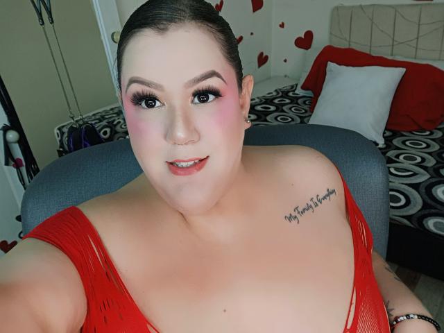 GiiannaMorrys has pale skin, pink blush, black eyeliner, red lipstick, and a tattoo on her right shoulder. She's wearing a red, mesh, low-cut top, showing her tits.