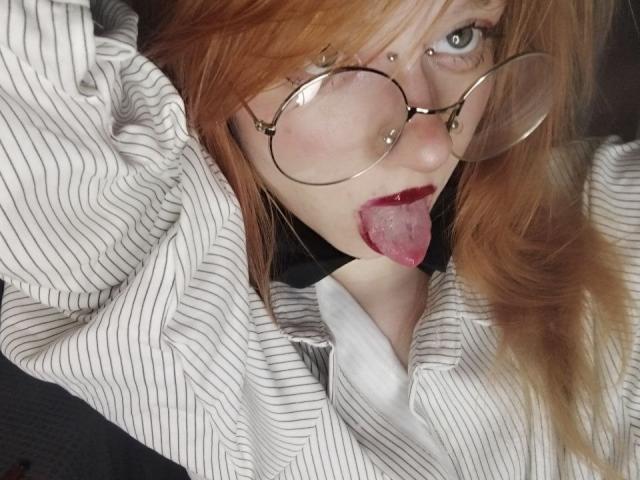 JamieLathrum with red hair, glasses, nose piercing, and black choker, sticking out tongue, wearing striped shirt, looking up, tongue out, glasses reflecting light, red lipstick.