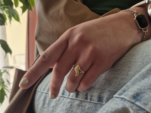 LoriEla's hand with a gold ring featuring a small, intricate design, wearing a bracelet, green nail polish, and light blue jeans, resting on a brown jacket.