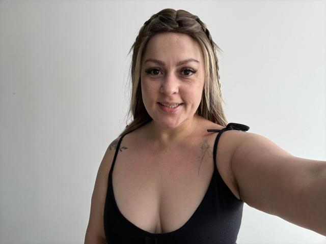 SamanthaGold, avec des cheveux bruns, un débardeur noir, de petits tatouages sur les épaules, souriante, tenant un téléphone, poitrine moyenne, peau claire, maquillage léger, fond blanc, selfie, décontractée, confiante..