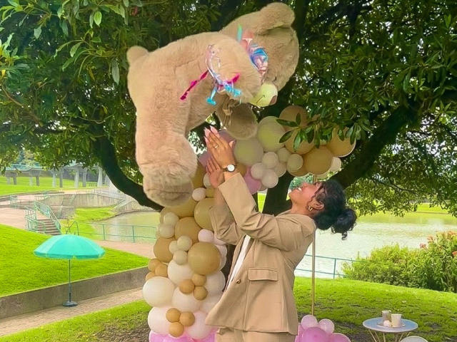 Sophie Sonya trägt einen beigen Anzug und hält einen riesigen, braunen Teddybären mit rosa und blauen Schleifen. Sie steht unter einem Baum mit Ballons, blickt nach oben und befindet sich in einem üppigen, grünen Park..