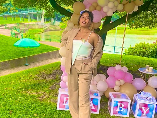 SophieSonyas with tan pants and white top, tan jacket, hands in pockets, pink and white balloons, pink and blue boxes, standing in grassy park, green umbrella in background.