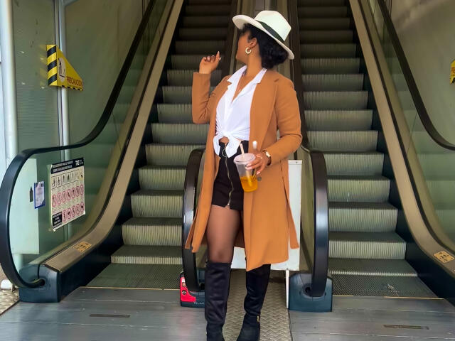 SophieSonyas, avec ses cheveux noirs bouclés, portant un manteau beige, une chemise blanche, un short noir, des bottes hautes jusqu'à la cuisse, un chapeau blanc, tenant une boisson orange et un sac à main noir, se tient devant des escalators..