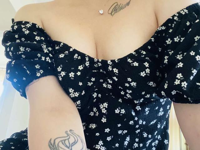 KarKutie's tits are showing in a black off-shoulder dress with white floral patterns. She has a tattoo on her arm and a necklace with a small heart.