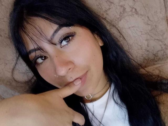LinaSpearce, avec de longs cheveux noirs, un piercing au nez, un piercing aux lèvres, un doigt dans la bouche, des yeux marron, une chemise blanche, un collier en or, et un joli sourire. Sans seins, sans fesses, sans vagin, piercing aux lèvres visible..