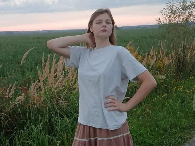 WilheminaBuchwalter, standing in grass, wearing a white tee and pink striped skirt, hand on hip, other arm behind head, looking serious, with a cloudy sunset in the background.