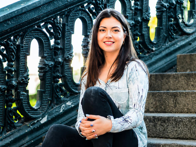 SamantaMais has long brown hair, smiling with white teeth, wearing a white patterned shirt and black pants, sitting on stone stairs with ornate black railings.