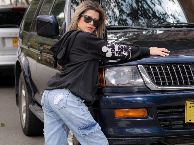 Sheyla Spooner está encostada em um SUV azul escuro, vestindo um moletom preto com desenhos brancos e calças jeans azuis rasgadas, com as nádegas levemente elevadas, e óculos de sol..