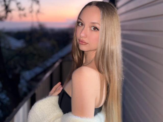 RandiMatute with long blonde hair, brown eyes, and pouty lips, wearing a black strap top, white fluffy sweater, looking over shoulder, sunset in background, standing on balcony.