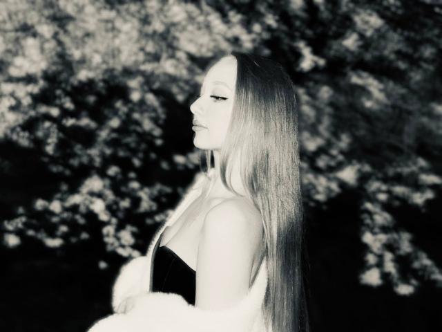 RandiMatute with long, straight, blonde hair, wearing a black, strapless top, and a fluffy white jacket, looking to the side with closed eyes, in a black-and-white photo.