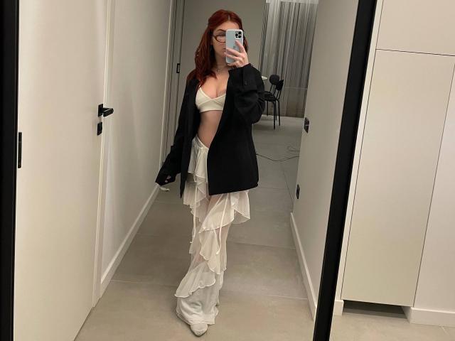 AlexXas with red hair, wearing a white bra, black jacket, and ruffled white skirt, taking a mirror selfie, showing her small tits and slim waist.
