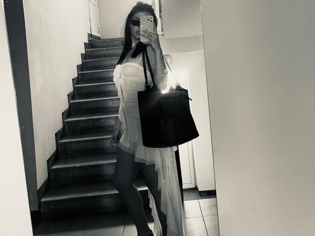 Foto in bianco e nero di AlexXas con i capelli lunghi, che indossa un abito bianco trasparente, tiene una borsa nera, è in piedi su un pavimento piastrellato e sta scattando un selfie allo specchio, mostrando il seno..
