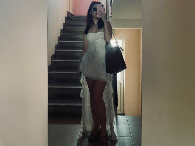 AlexXas in a white, strapless, ruffled dress with a long back, holding a black bag, standing on dark tiles, taking a mirror selfie with visible tits and brown sandals.