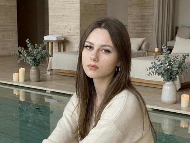 KimberlyZampieri has long brown hair, subtle makeup, and is wearing a beige sweater. She's sitting by a pool with candles and greenery in the background. Boobs are not visible.