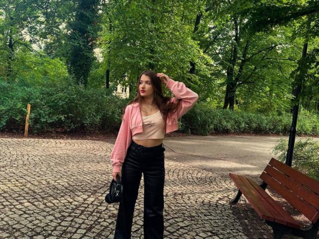 MisscharyNessy with brown hair, pink jacket, white top, black pants, holding camera, standing on cobblestone path, hand in hair, wooden bench to the right, green trees in background.