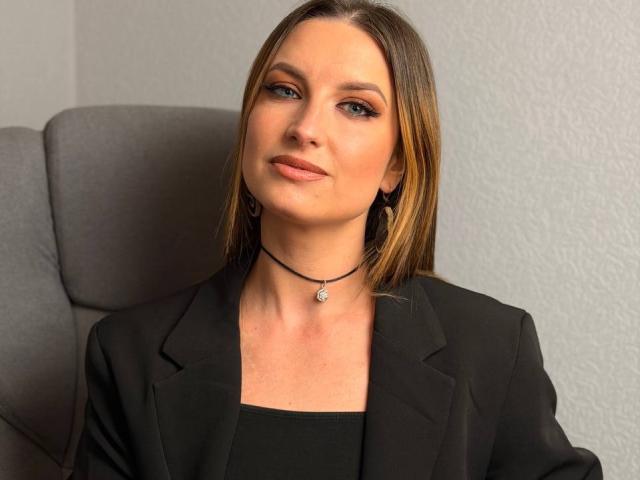 Milagres has medium-length brown hair, blue eyes, and is wearing a black blazer with a black choker and small pendant. She's sitting in a gray office chair.
