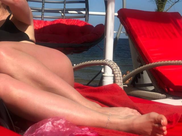 Milagres' ass and legs on a red towel, wicker chair, wearing a black bikini, ankle bracelet, pink bag, sunny day, blue water in background.