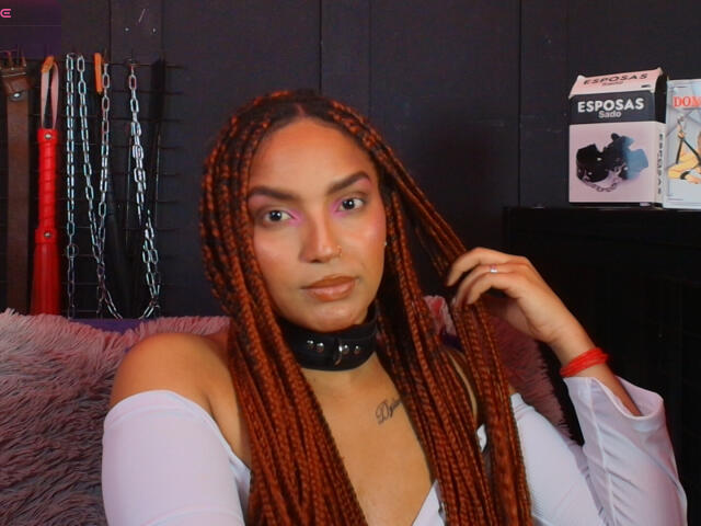 BrianaPratt with long braided red hair, wearing a white off-shoulder top, black collar, and nose ring, looking at camera, hand in hair, red bracelet, soft smile.