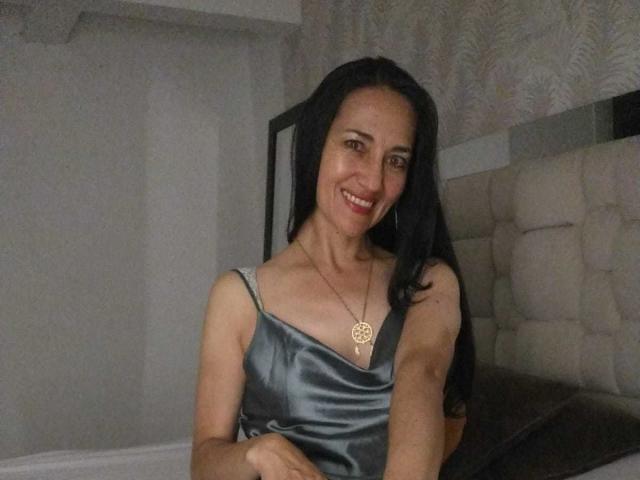 MiaMott with black hair, smiling, wearing a shiny gray satin dress, gold necklace with a circular pendant, showing her tits, in a bedroom with a tufted headboard.