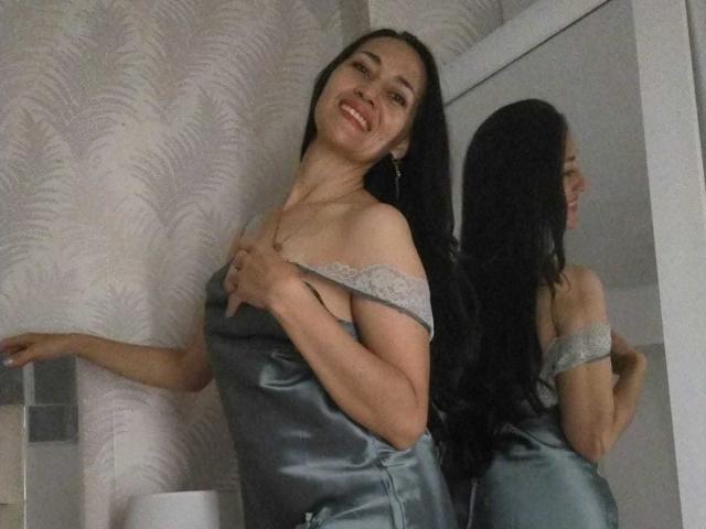 MiaMott with long black hair, smiling, wearing a shiny gray satin dress with lace off-shoulder, her tits slightly visible, standing in front of a mirror.
